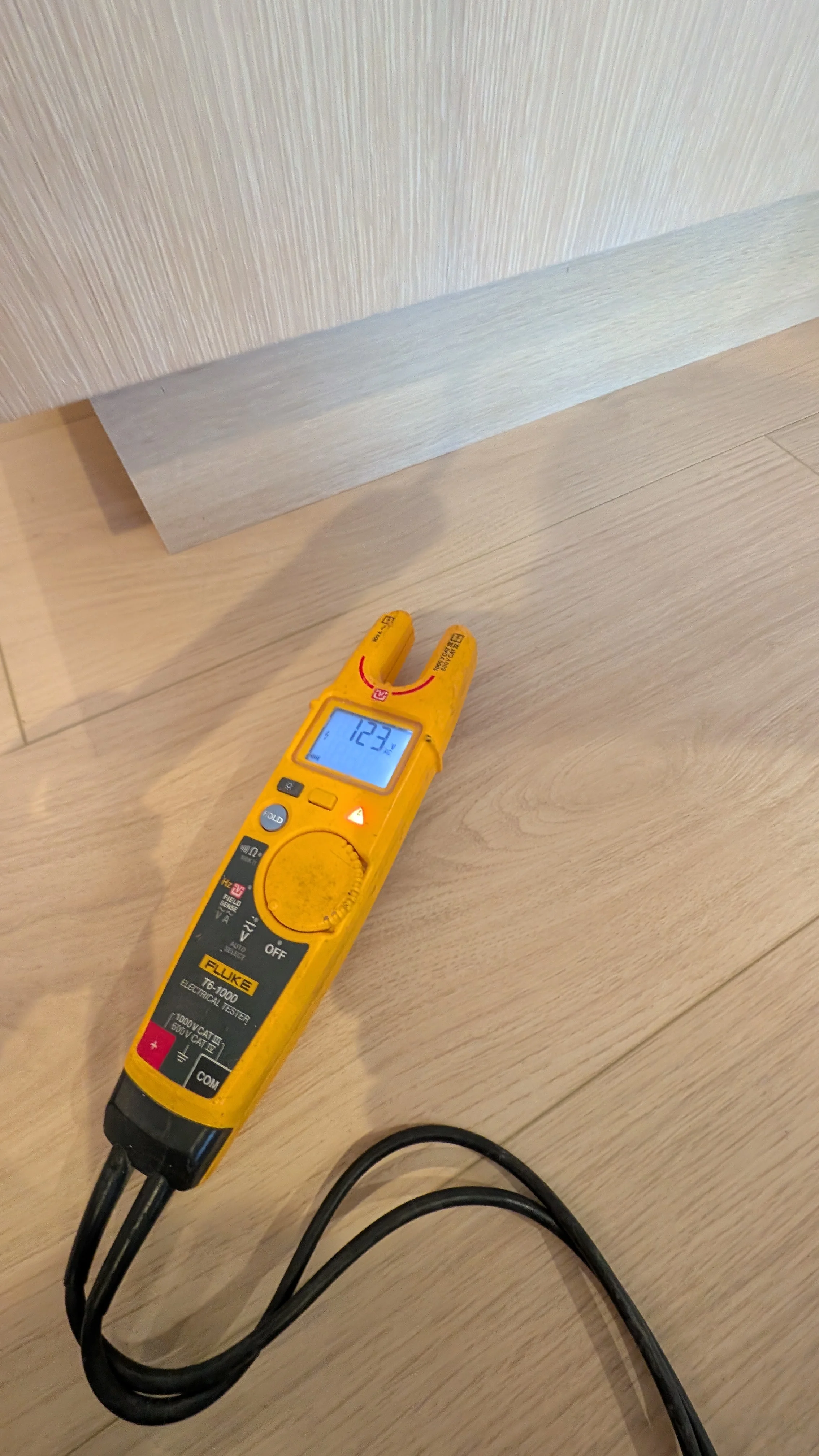 Digital clamp meter displaying voltage reading on light wood flooring during electrical inspection