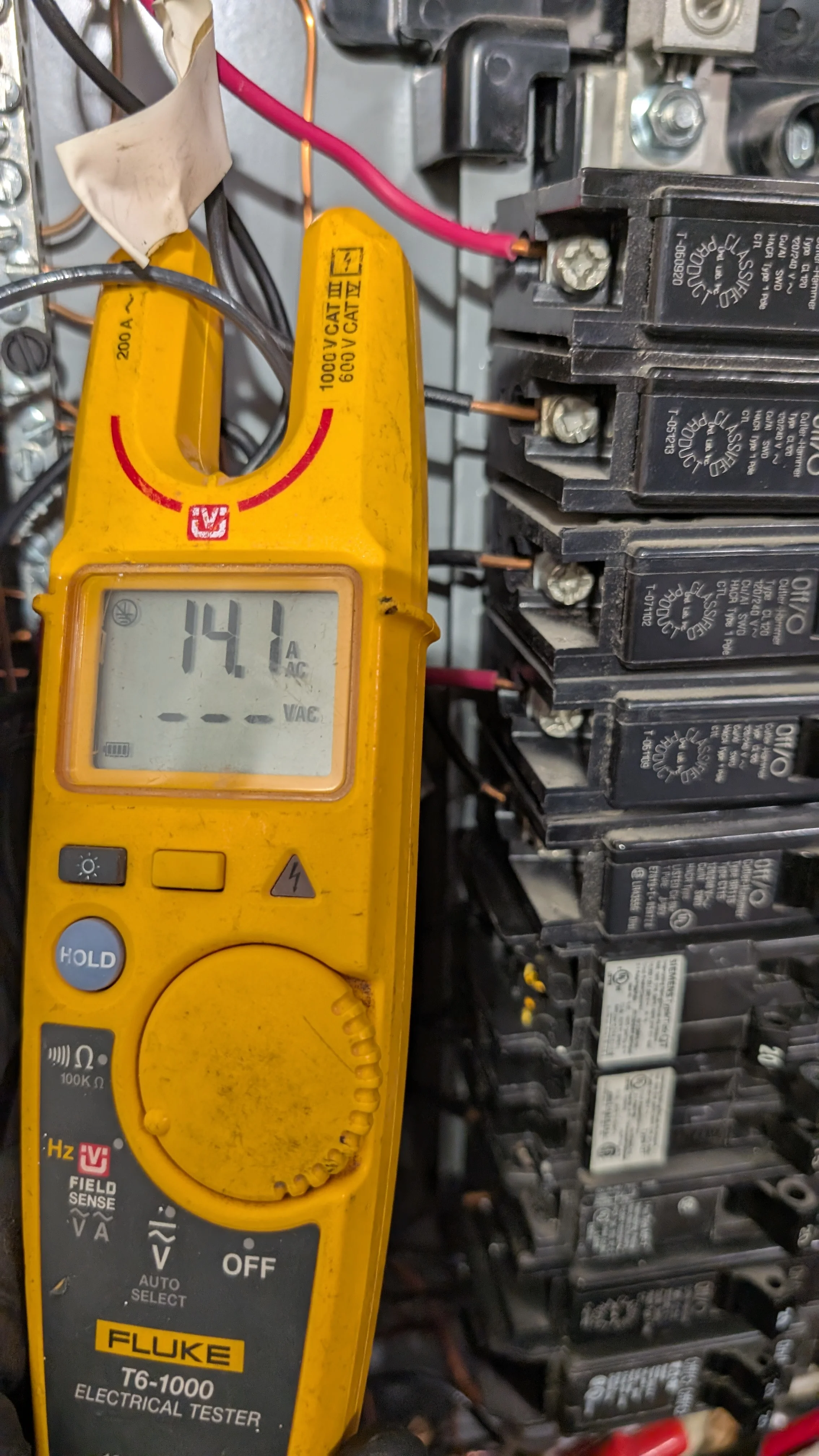 Yellow Fluke clamp meter measuring 14.1 amps at residential electrical panel with multiple black breakers